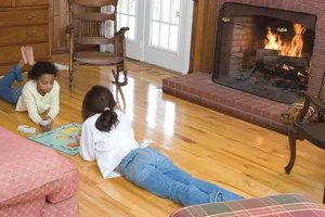 Two children lying on a light hardwood floor in front of a lit brick fireplace